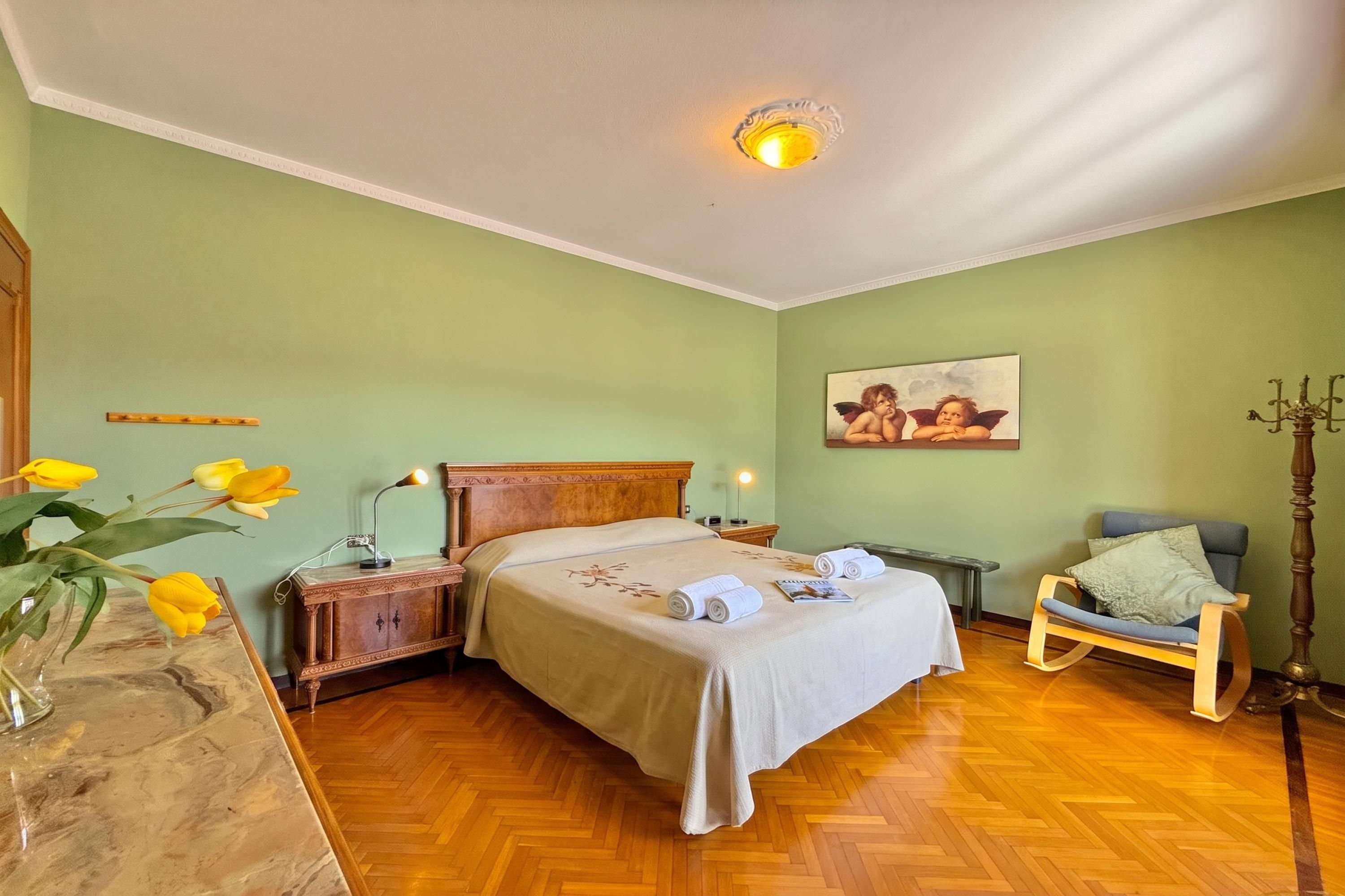 A hotel room with a bed, bedside tables, a chair, a painting, and a vase with flowers.
