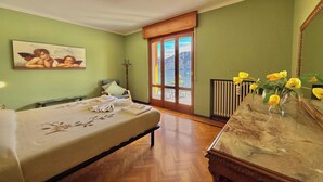 A hotel room with a bed, a wooden dresser, a painting, a window with a view of mountains, and a vase with flowers. - Isola Vista - Terrazzo (Sala Comacina)