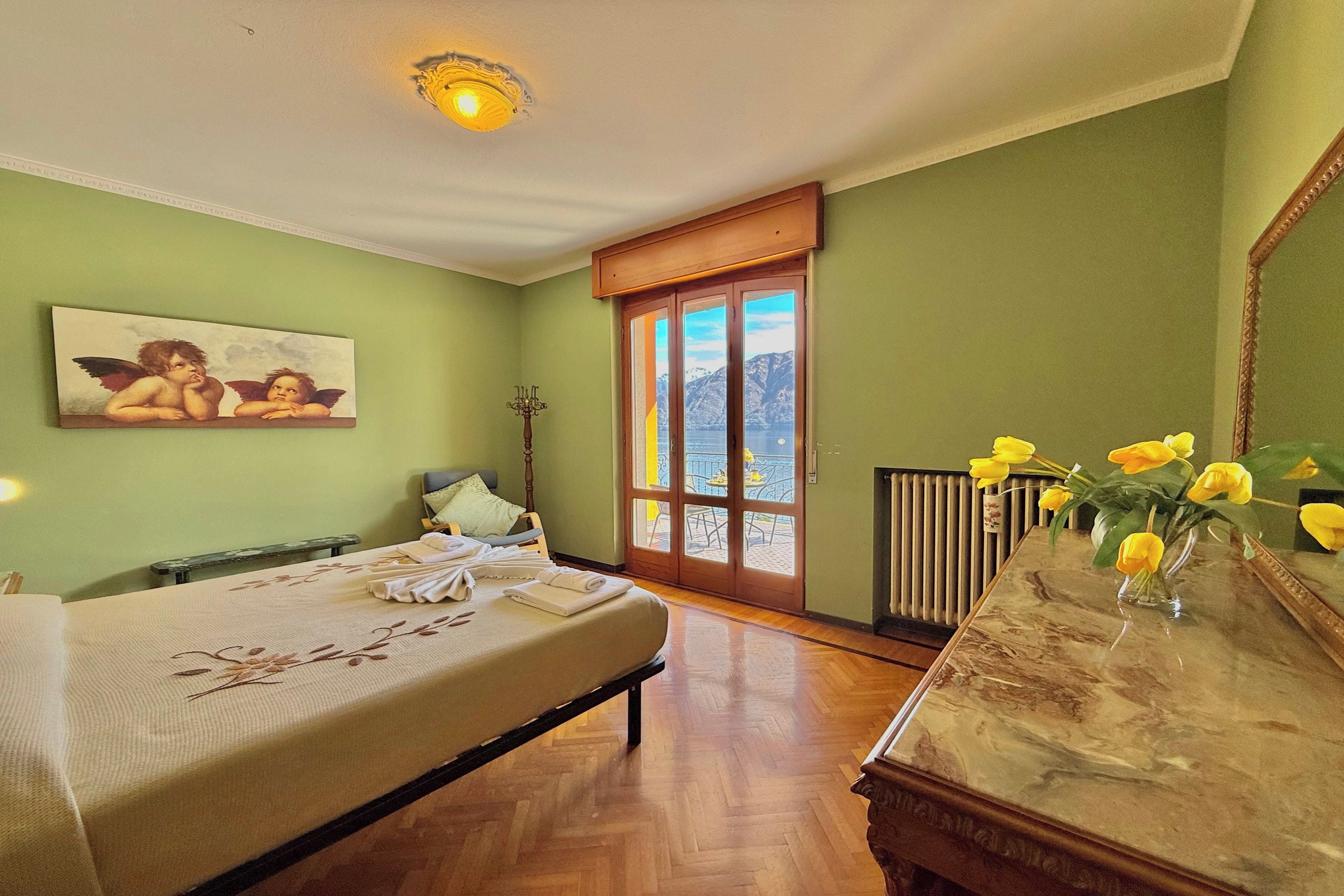 A hotel room with a bed, a wooden dresser, a painting, a window with a view of mountains, and a vase with flowers.