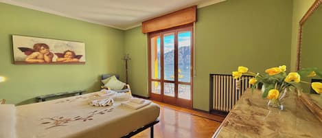 A hotel room with a bed, a wooden dresser, a painting, a window with a view of mountains, and a vase with flowers.