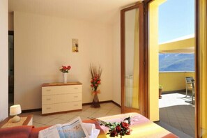 A room with a bed, a dresser, a vase with flowers, and a view of a balcony with a chair.