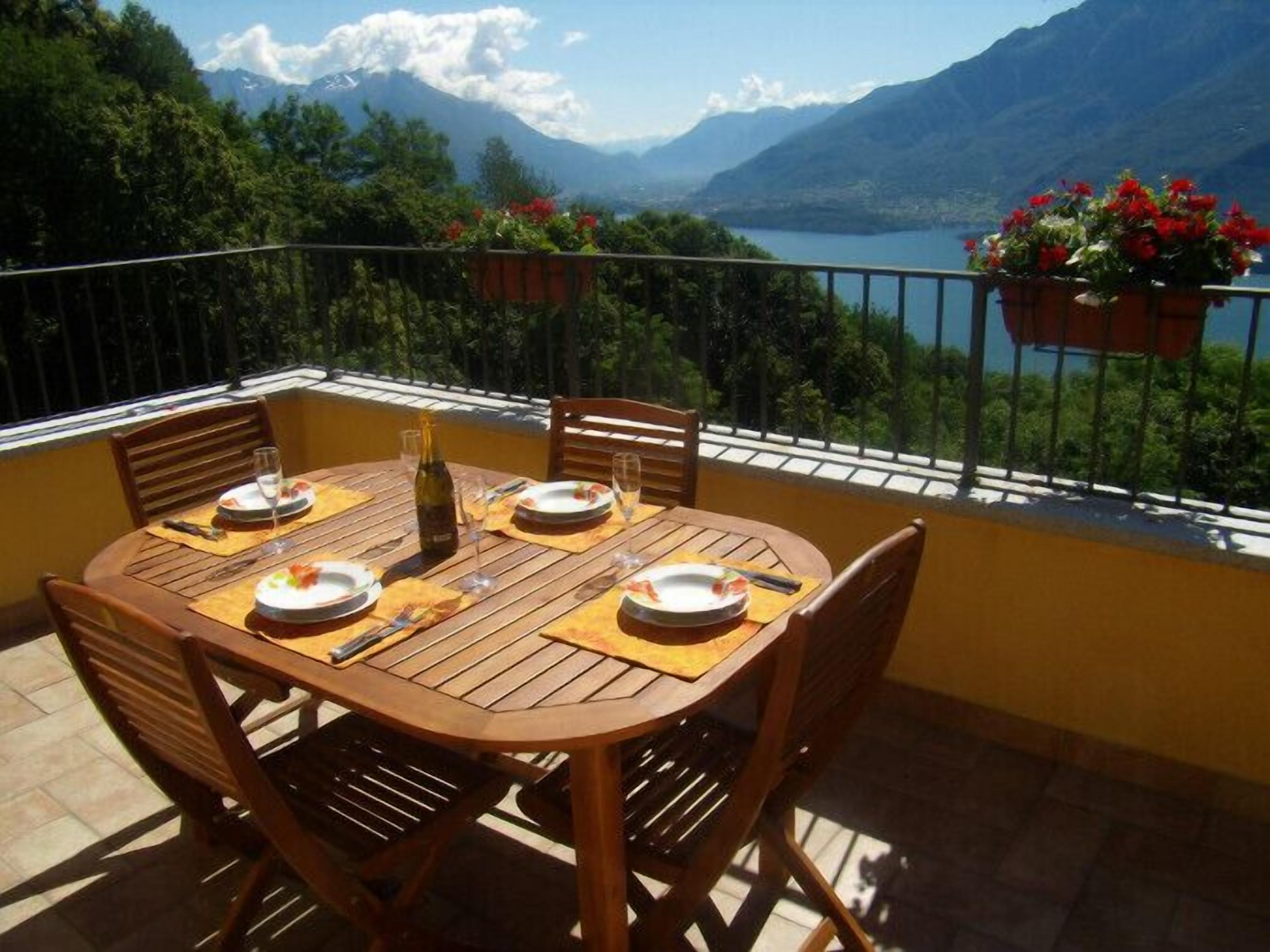 A wooden table set for four with plates, cutlery, and a bottle of wine, overlooking a lake and mountains.