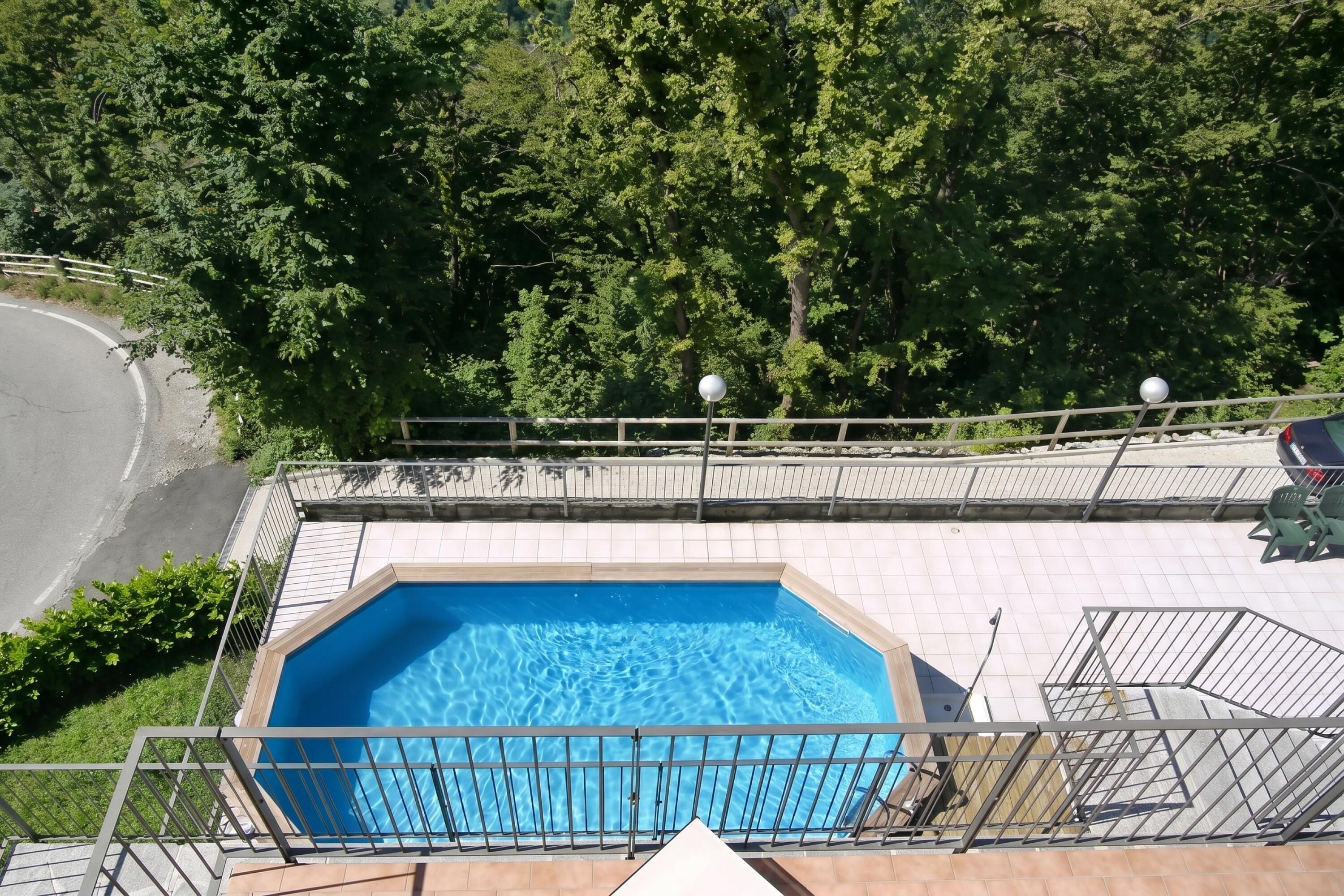 A swimming pool with a tiled deck, surrounded by a metal fence and trees.