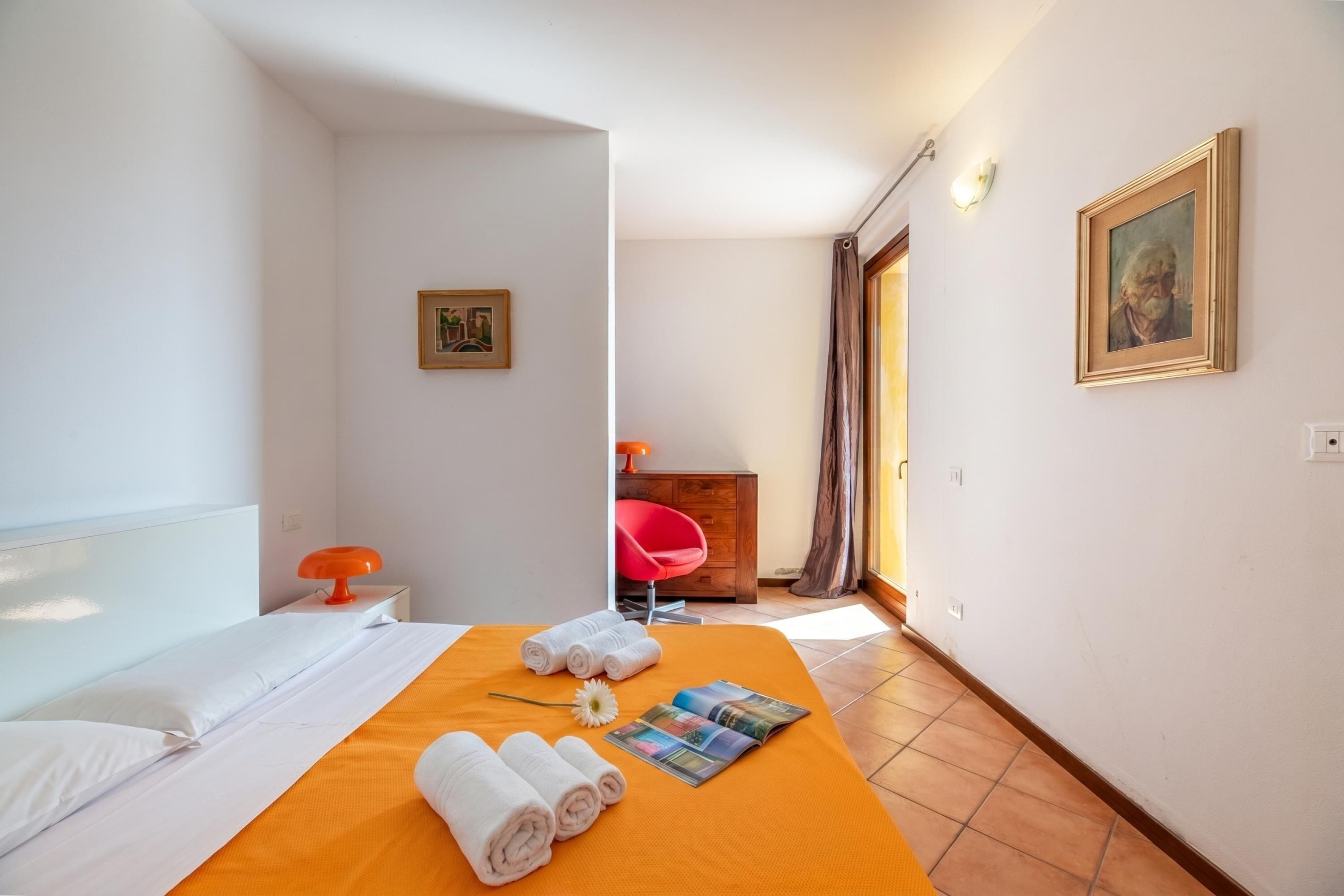 A hotel room with a bed, a nightstand, a red chair, a painting, and a door leading to another room.