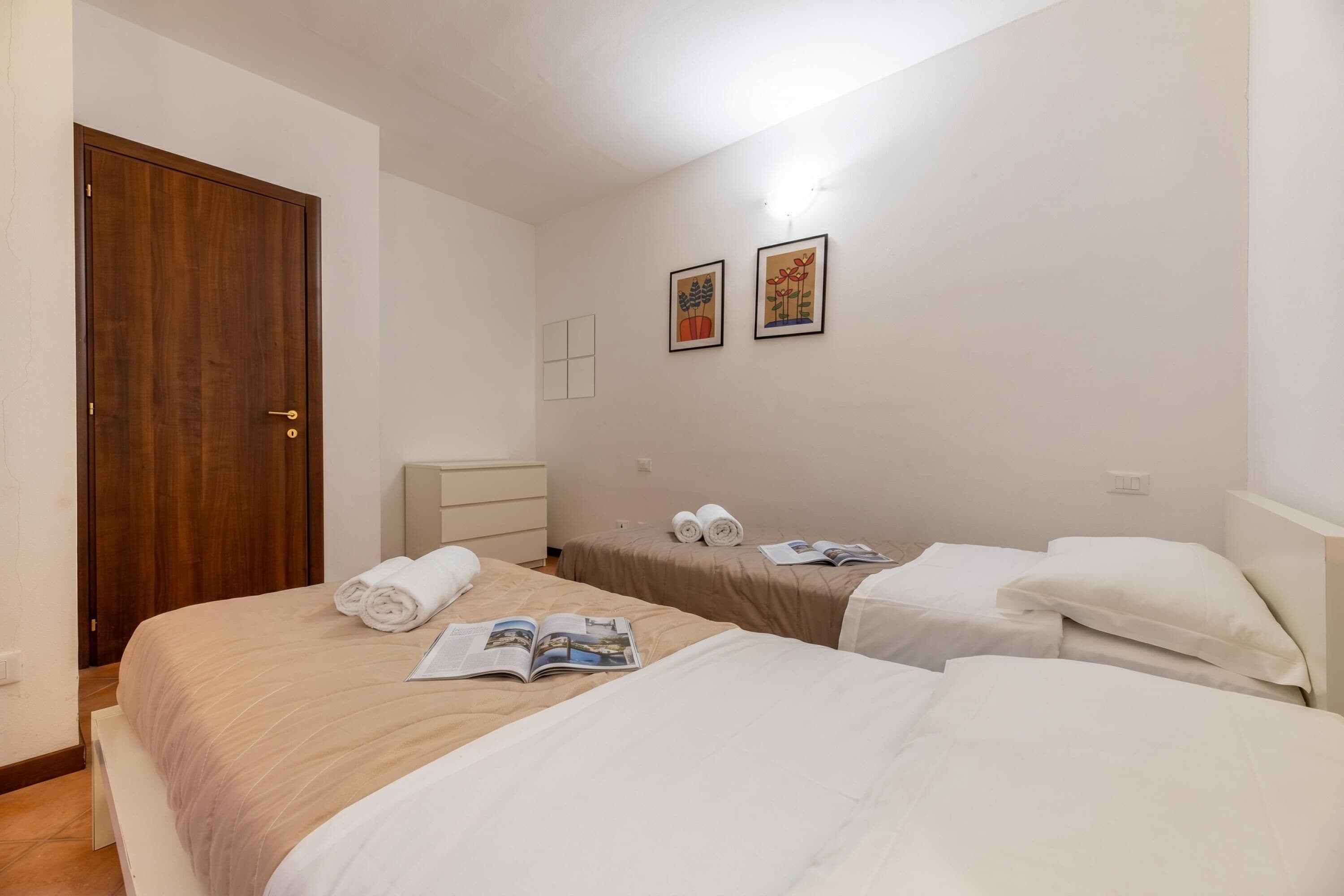 Une chambre d’hôtel avec deux lits, une porte en bois, une table de chevet blanche et deux tableaux encadrés au mur.