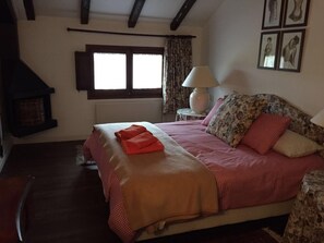 A bedroom with a bed, a lamp, a fireplace, and framed pictures on the wall.