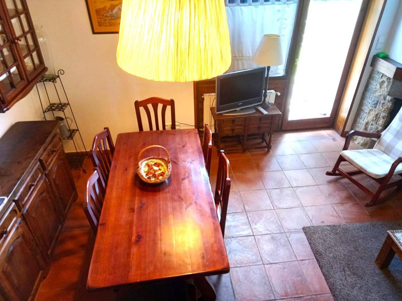 A dining area with a wooden table, chairs, a basket of fruit, a television, and a lamp.