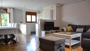 Un salon moderne avec un canapé, un pouf et une table basse en verre. Il y a également un coin cuisine avec des placards et un téléviseur.