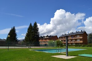 A swimming pool with a fence, a tennis court, and a building with balconies.