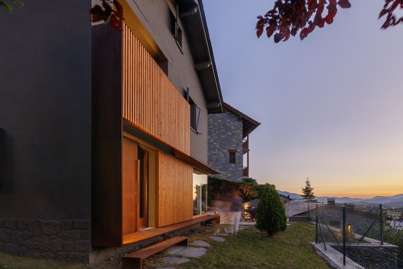 Uma casa moderna de dois andares com fachada de pedra, ripas de madeira na fachada externa, um deck de madeira e vista para as montanhas.