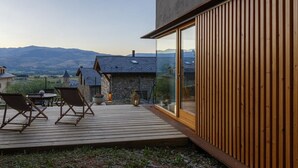 A wooden deck with chairs, a stone building, and a mountain view.