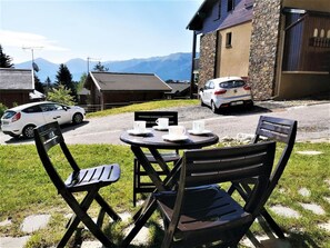 En terrasse med udendørs møbler, en hvid bil og en stengul bygning med udsigt til et bjerg.