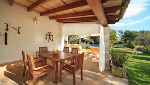 Un patio exterior con mobiliario de madera, un dosel blanco y vistas a una piscina y jardín.