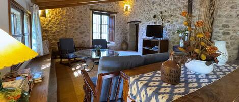A rustic living room with stone walls, a wooden ceiling, a sofa, a TV, and a table with a patterned tablecloth.