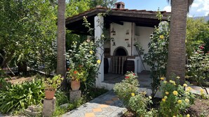 A rustic building with a tiled roof, a stone archway, and a garden with various plants and flowers. - Maros Spitaki (Varipetro)