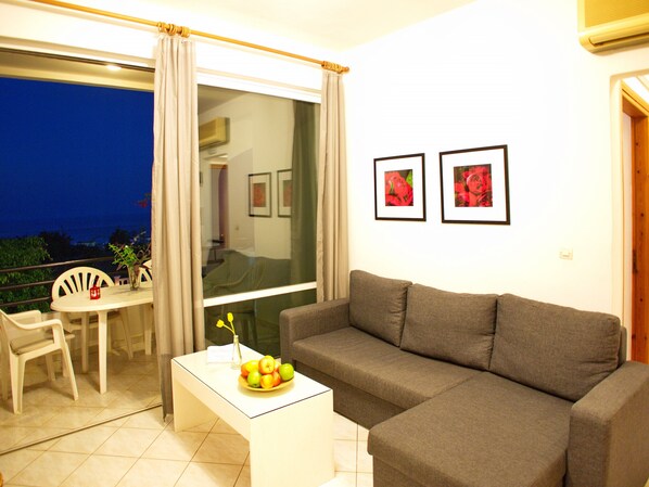 A living room with a grey sofa, a small white table with a fruit bowl, and a balcony with chairs.