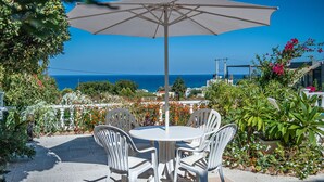 En terrasse med et rundt bord og stole, en stor parasol og udsigt til havet.