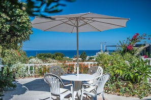 Eine Terrasse mit rundem Tisch und Stühlen, einem großen Sonnenschirm und Blick auf das Meer.
