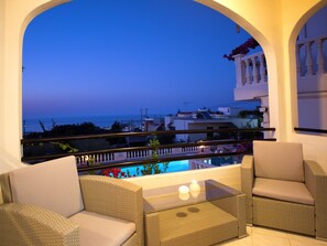 A balcony with wicker furniture, a glass table, and a view of the sea and buildings.