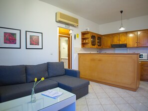 A living room with a grey sofa, a glass coffee table with a book, a vase with a flower, and a kitchen area with wooden cabinets and a microwave. - Rouladina Apt 3 (Stalos)