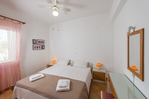 Une chambre à coucher avec un lit, deux lampes de chevet, un ventilateur de plafond, une fenêtre avec un rideau rose et un miroir fixé au mur.