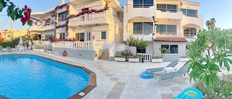 A poolside area with a clear blue pool, white tiled flooring, and a white building with balconies adorned with pink flowers.