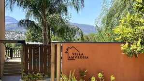 Entrada de una villa con un letrero que dice "Villa Amvrosia", una puerta de madera, plantas en macetas y una vista de la montaña y el pueblo al fondo.