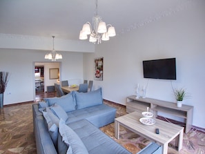 A modern living room with a sectional sofa, a dining area, a flat-screen TV, and a chandelier.