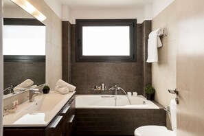 A modern bathroom with a large bathtub, a double sink, and a toilet. There is a window above the bathtub and a towel rack on the wall. - Villa Anemi (Sternes)