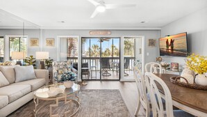 Un salon spacieux avec un canapé, une table basse et un coin repas. Une pancarte indiquant « MAISON DE PLAGE » est placée au-dessus de l’entrée.