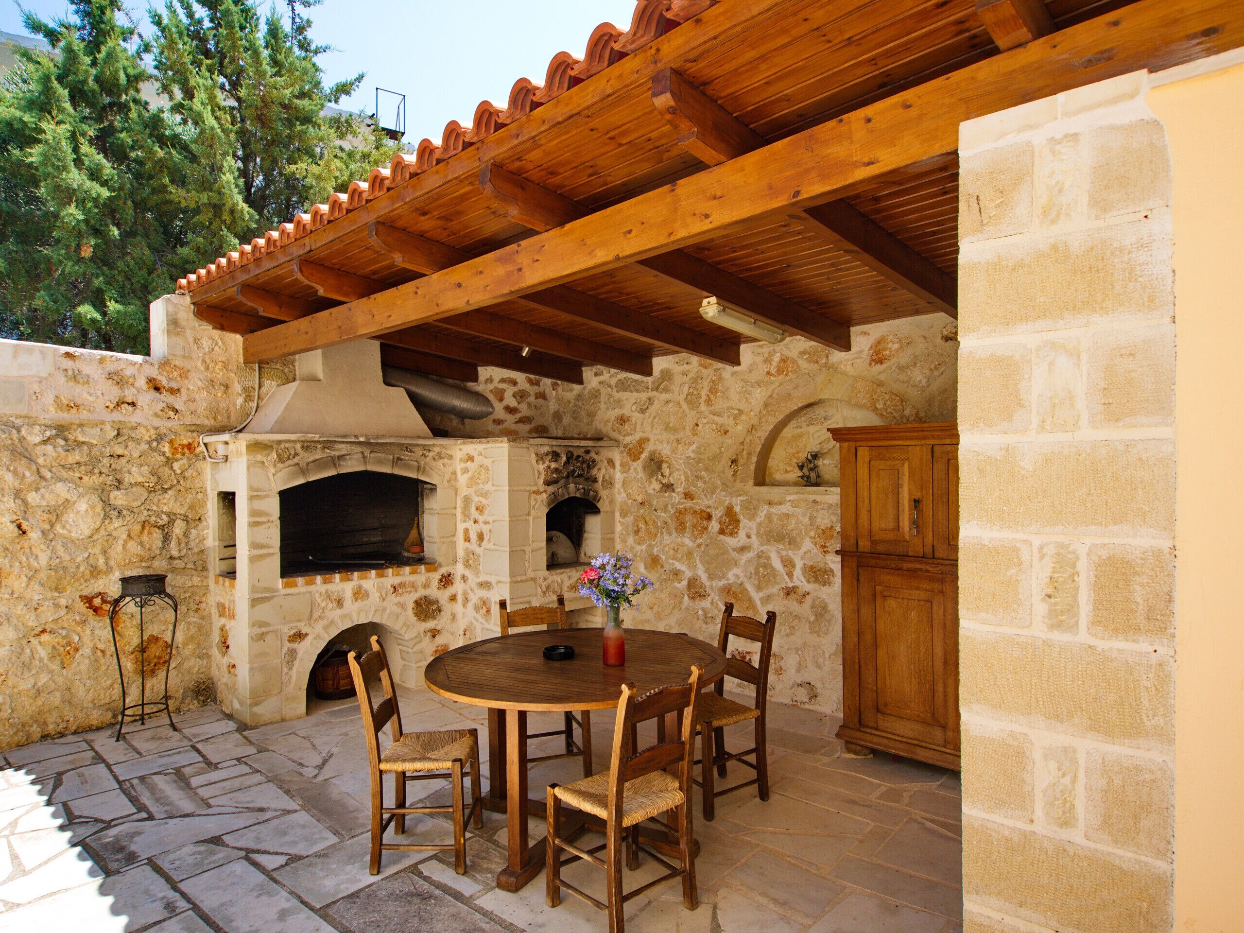 An outdoor patio with a stone wall, wooden table, chairs, and a pizza oven.