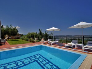 A swimming pool with lounge chairs and umbrellas, surrounded by a stone wall and greenery, overlooking a scenic landscape. - Villa Artemis (Vrises)