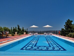 Piscina con sillas de descanso y sombrillas, rodeada de un muro de piedra y vegetación.