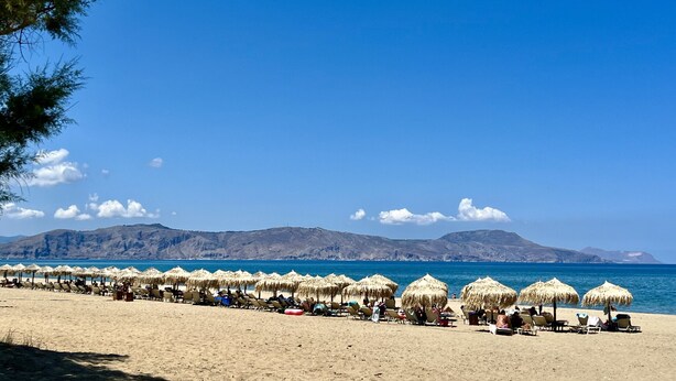 Ein Strand mit Liegen und strohgedeckten Sonnenschirmen, ein klarer blauer Himmel und ferne Berge.