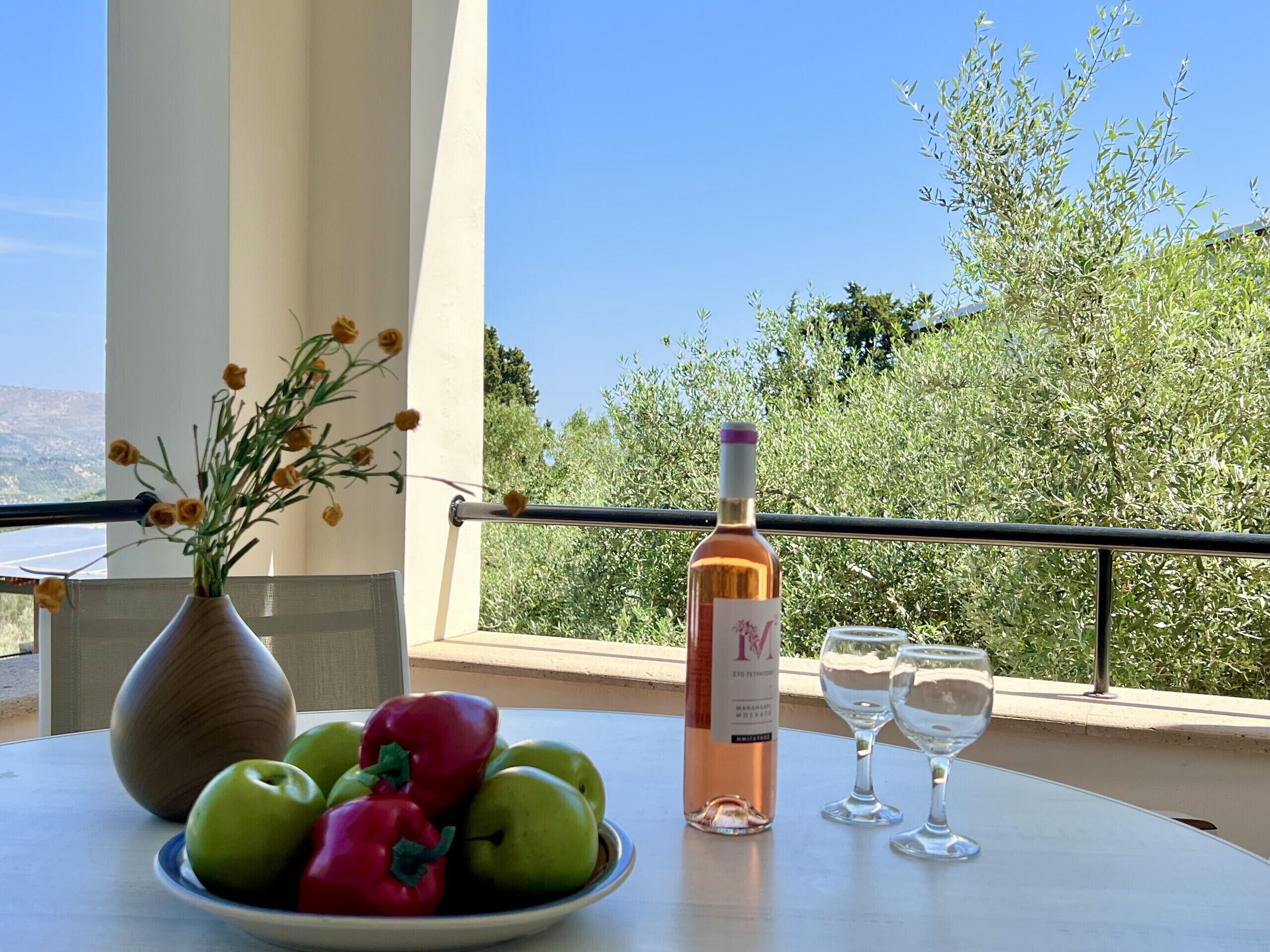 Et bord med en vinflaske, to glas, en skål med æbler og peberfrugter, og en vase med blomster på en balkon med udsigt til grøn vegetation og en klar himmel.