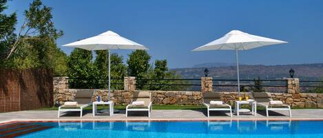 A pool area with lounge chairs and umbrellas, surrounded by stone walls and a view of the landscape.