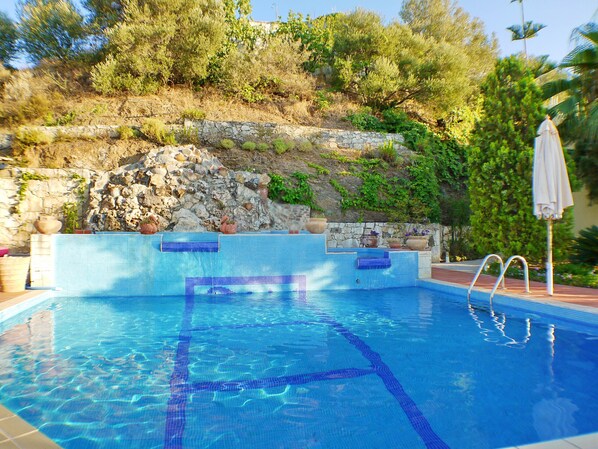 A swimming pool with clear blue water, stone steps leading into the pool, and a stone wall with greenery behind it.