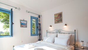 A bedroom with a bed, a nightstand, a window with curtains, a wall-mounted picture, and a wall-mounted light fixture. - Villa Clio (Stalos)