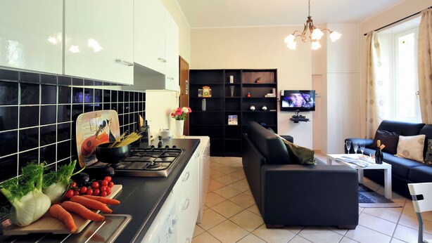 A modern kitchen with a stove, sink, and counter, a black sofa, a television, and a bookshelf. - Villa Isella - Mansarda (Carlazzo)