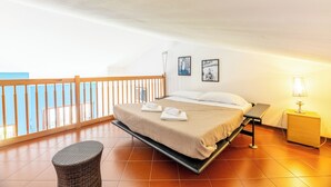 A bedroom with a bed, a nightstand, a lamp, a small table, a wicker chair, and a balcony with a railing. - Balcone di Noa e Stella (Menaggio)