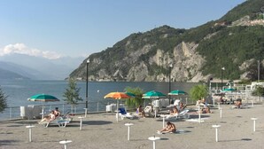 A beach with sun loungers and umbrellas, a rocky hillside in the background, and a clear view of the water. - Alla Vigna Panoramica (Perledo)