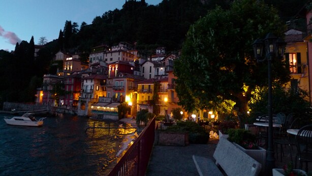 A waterfront town with buildings along the river, a bench, and a boat docked on the water. - Alla Vigna Panoramica (Perledo)