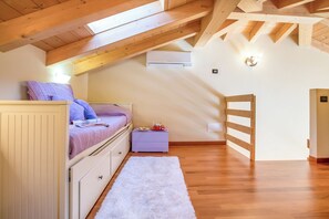 Quarto com teto inclinado, vigas de madeira, cama com almofadas, mesa de cabeceira, tapete branco e uma porta.