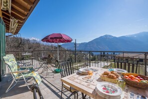 Terraço com mesa posta para refeições, cadeiras, um guarda-sol vermelho e vista para a montanha.