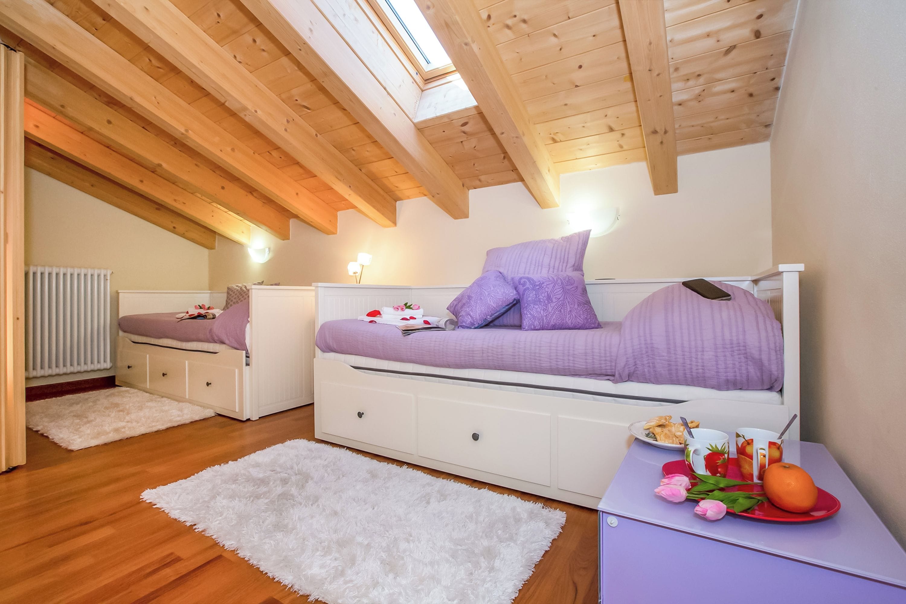 Una camera da letto con soffitto spiovente, travi in legno, un letto bianco con biancheria viola, un comodino con un piatto di frutta e un tappeto bianco e soffice.