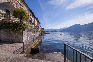 A waterfront view with buildings, a calm lake, and mountains in the background. - Colonno Panoramica (Colonno)