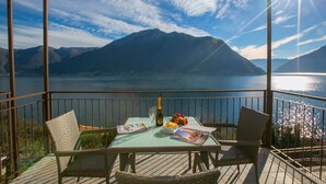 A balcony with wicker chairs, a table with a bottle, glasses, and a bowl of fruit, overlooking a lake and mountains. - Colonno Panoramica (Colonno)