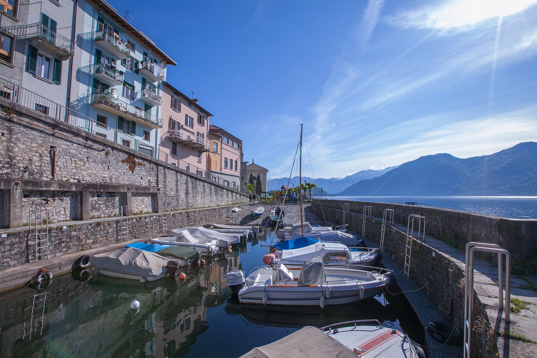 Un port de plaisance où des bateaux sont amarrés le long d’un quai en pierre, offrant une vue sur une étendue d’eau avec des montagnes en arrière-plan.