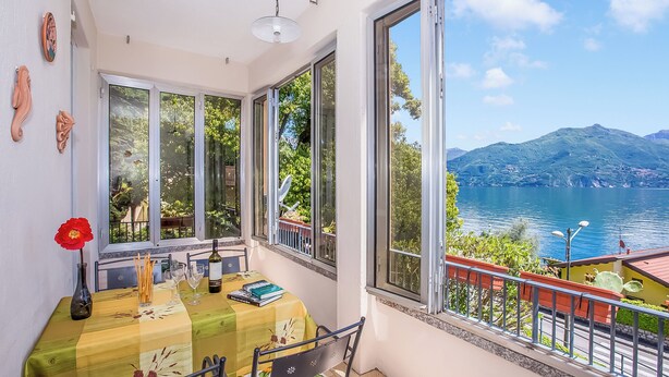Un balcon avec une table, des chaises, un vase contenant une fleur rouge, et une vue sur un lac et des montagnes.