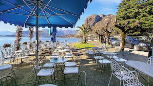 Un café en plein air avec des tables et des chaises, un parasol bleu et une vue sur la mer et les montagnes.